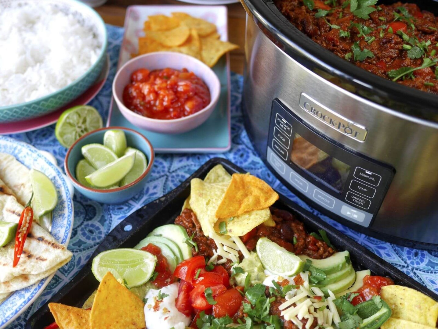 Ein Tisch mit Chili, Reis, Tortilla-Chips, Salsa, Limetten und anderen Belägen. Rechts ein Schongarer.