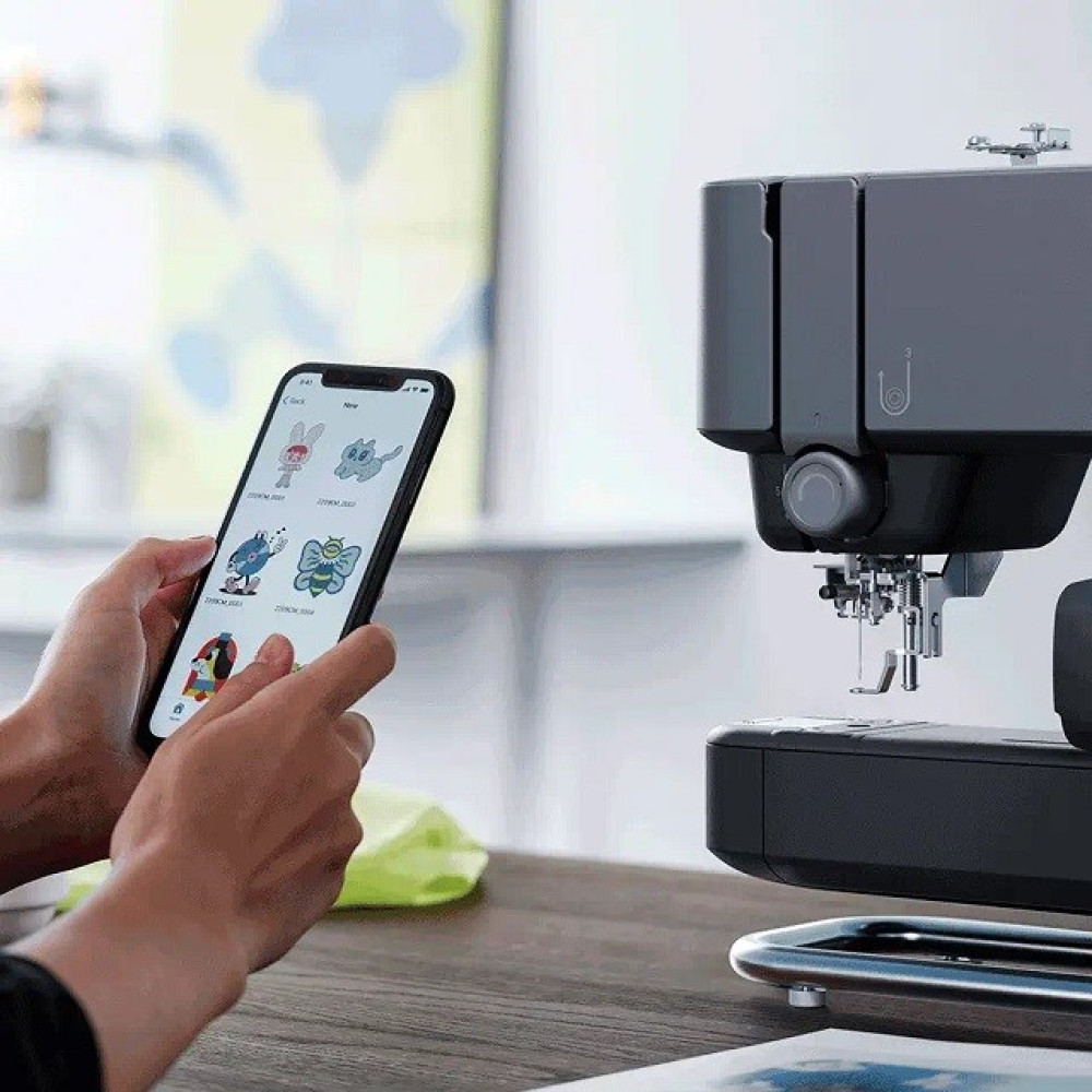 Ręce trzymają telefon z wzorami haftu, obok maszyny do szycia na drewnianym stole. Zielona szmatka leży na stole.