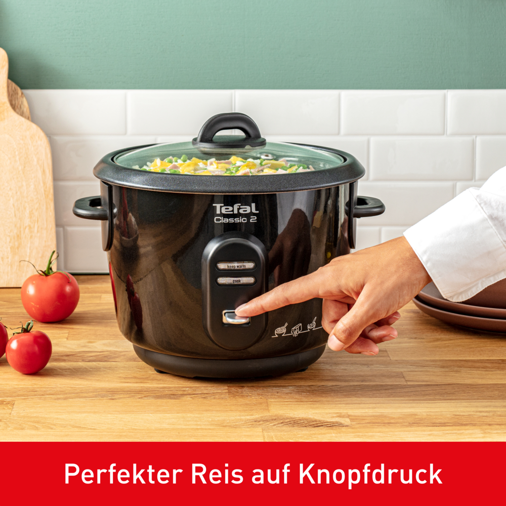 Eine Hand drückt die 'Cook'-Taste an einem schwarzen Tefal-Reiskocher, der mit Gemüse gefüllt ist. Tomaten und ein Schneidebrett im Hintergrund.