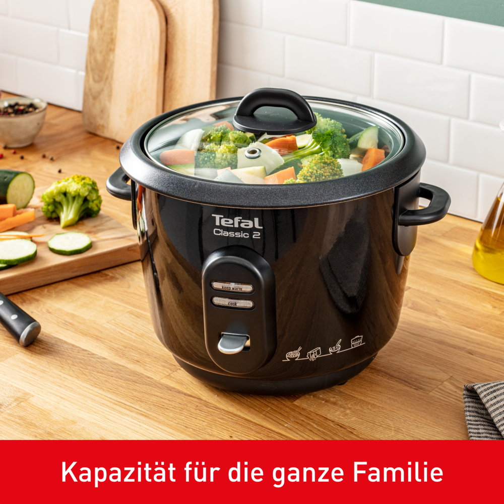 Ein schwarzer Tefal Classic 2 Reiskocher mit Gemüse. Gehacktes Gemüse liegt auf einem Holzbrett.