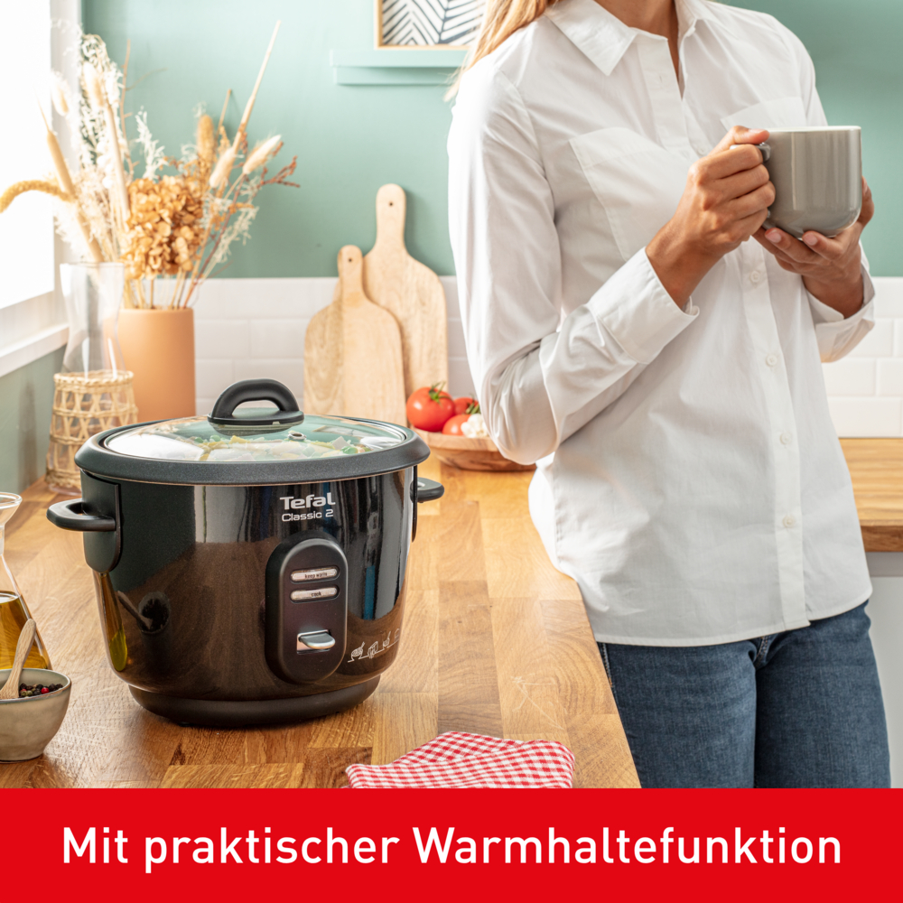 Eine Frau mit einer Tasse steht neben einem Reiskocher. Die Küche hat Holzarbeitsplatten, Schneidebretter und ein rot kariertes Tuch.