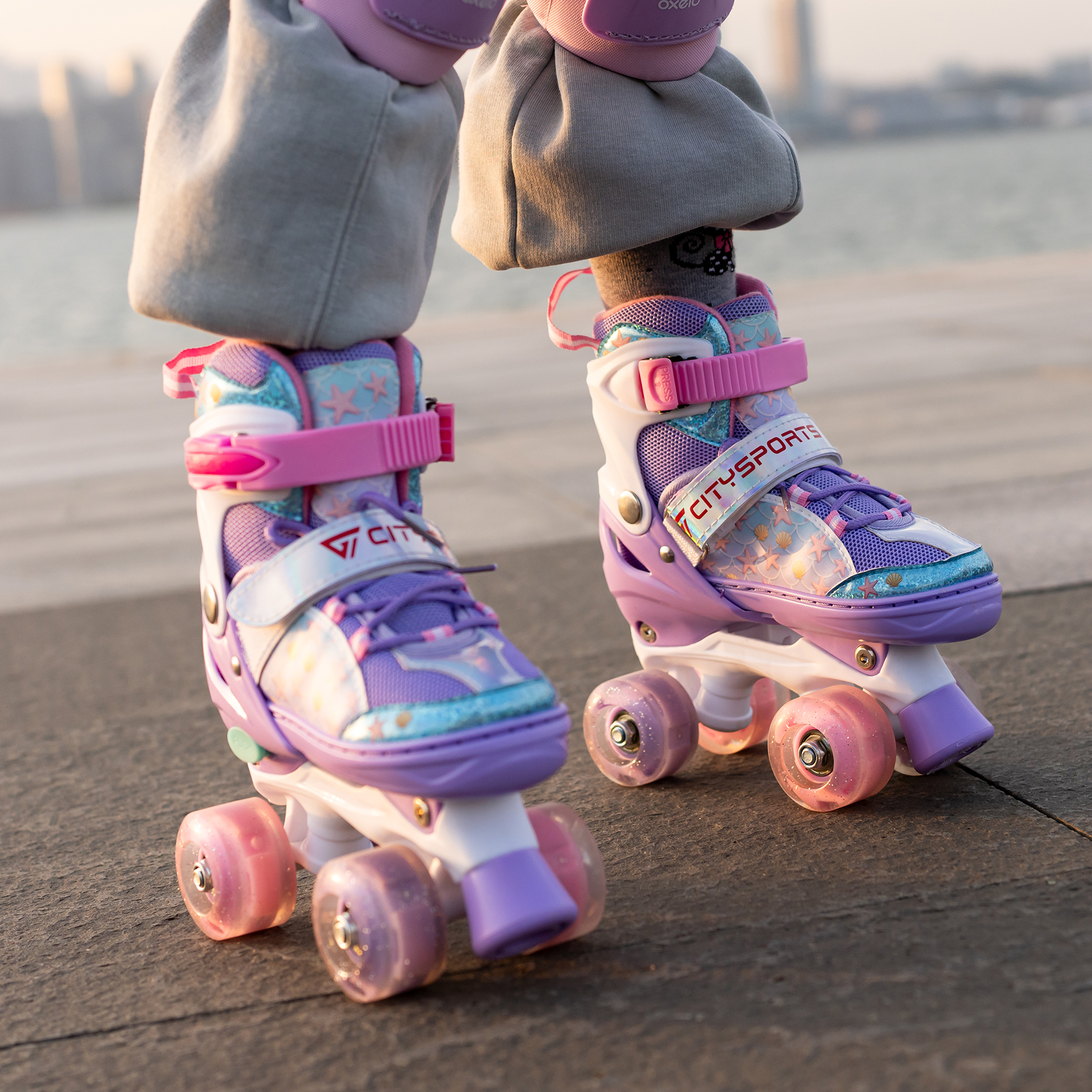 Piedi di bambino con pattini a rotelle viola e rosa. Ginocchiere. Sfondo: acqua e skyline cittadino.