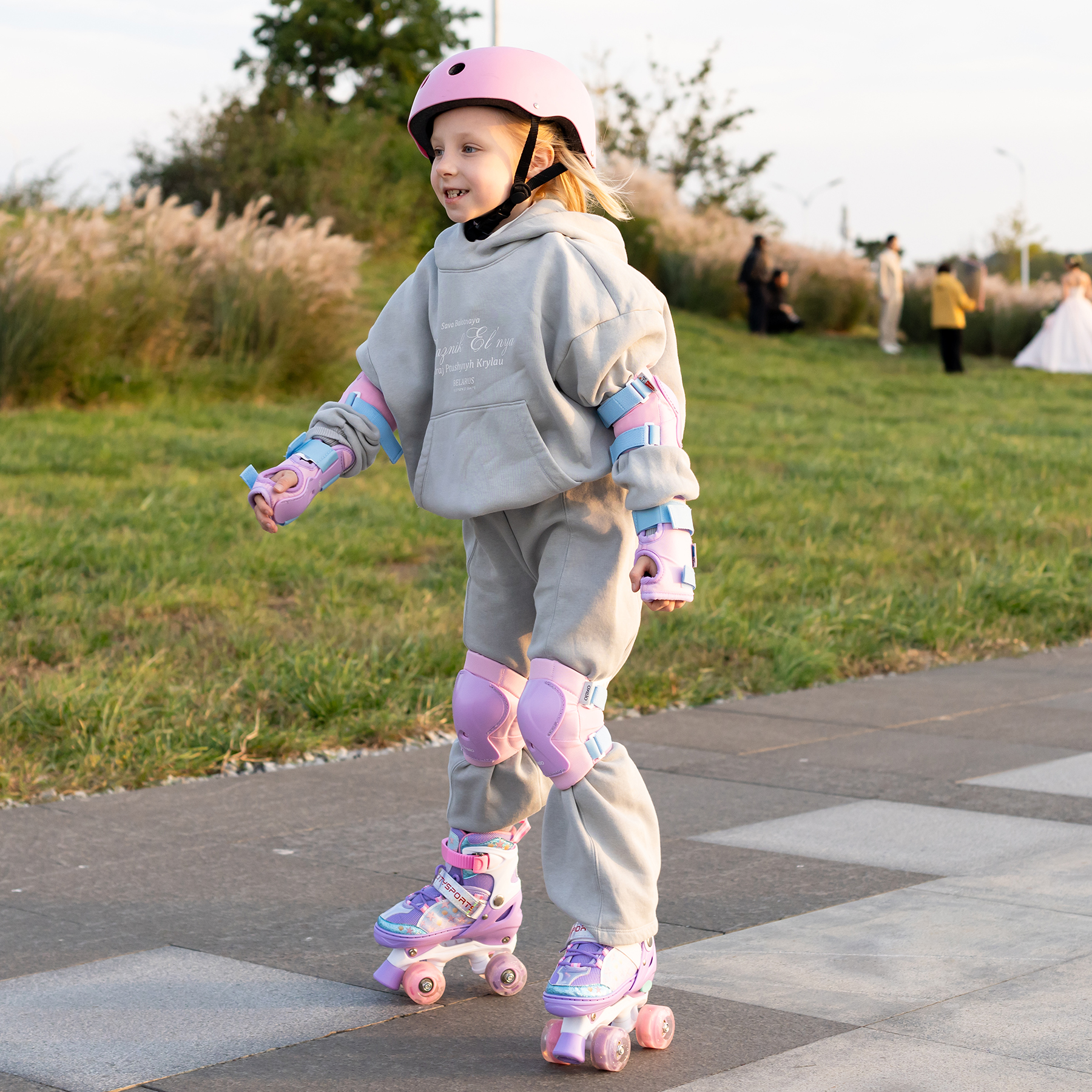 Una ragazza con casco rosa e protezioni pattina su una superficie pavimentata. Lo sfondo presenta erba, cespugli e persone.