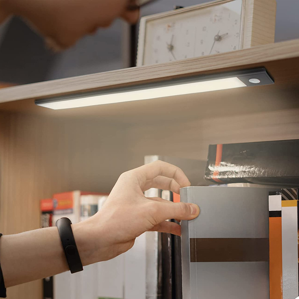 Eine Hand greift nach einem Buch im Regal. Oben ist ein Licht. Im Hintergrund sind eine Person und eine Uhr zu sehen.