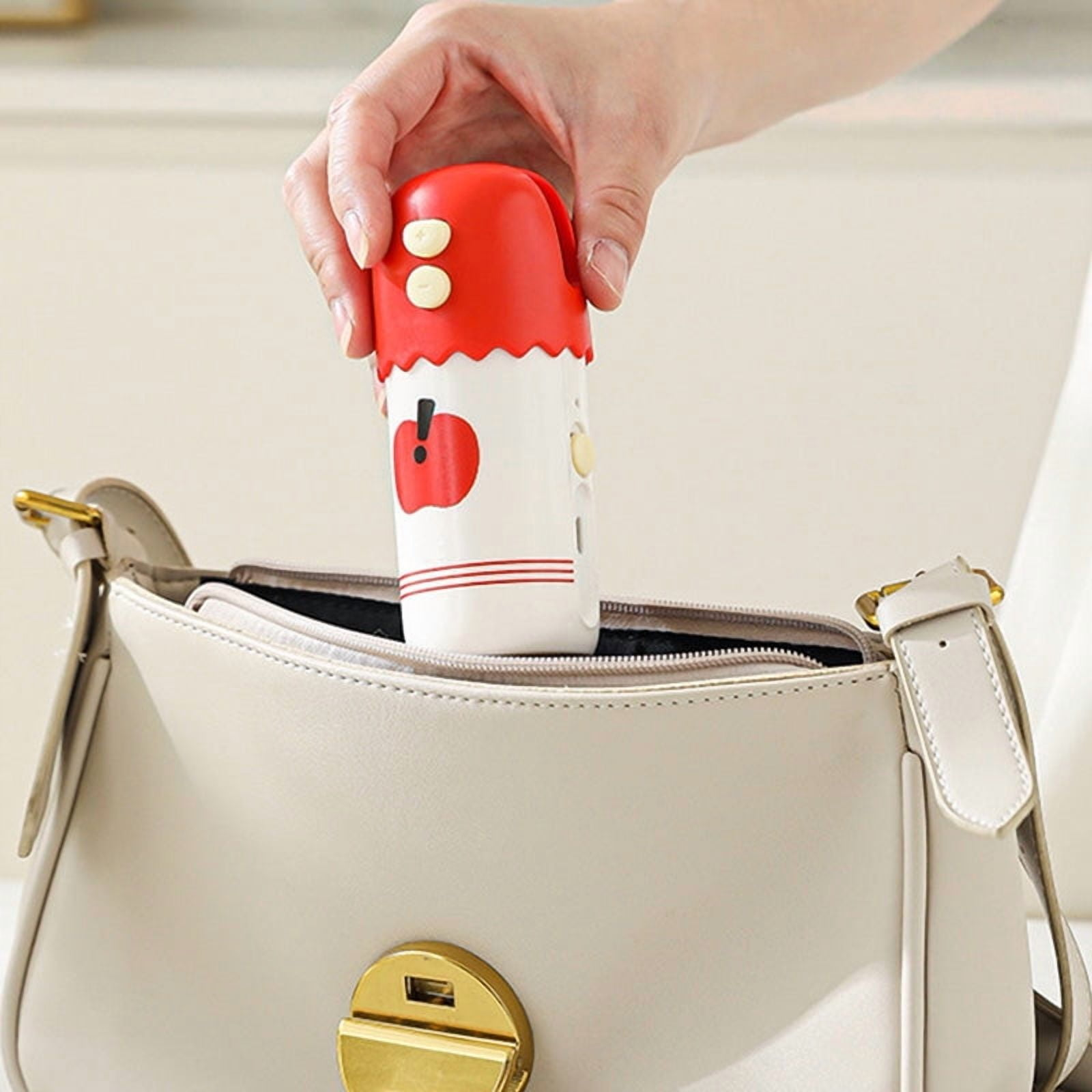 Eine Hand hält einen rotbedeckten Behälter mit Apfelmuster und legt ihn in eine beigefarbene Handtasche.