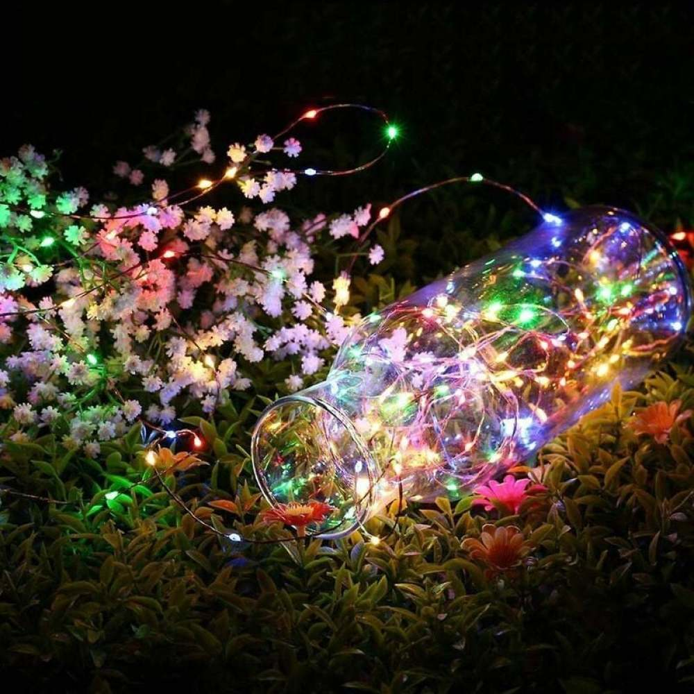 Ein Glasgefäß liegt im Gras und ist mit bunten Lichtern gefüllt. Weiße und orangefarbene Blumen umgeben es.