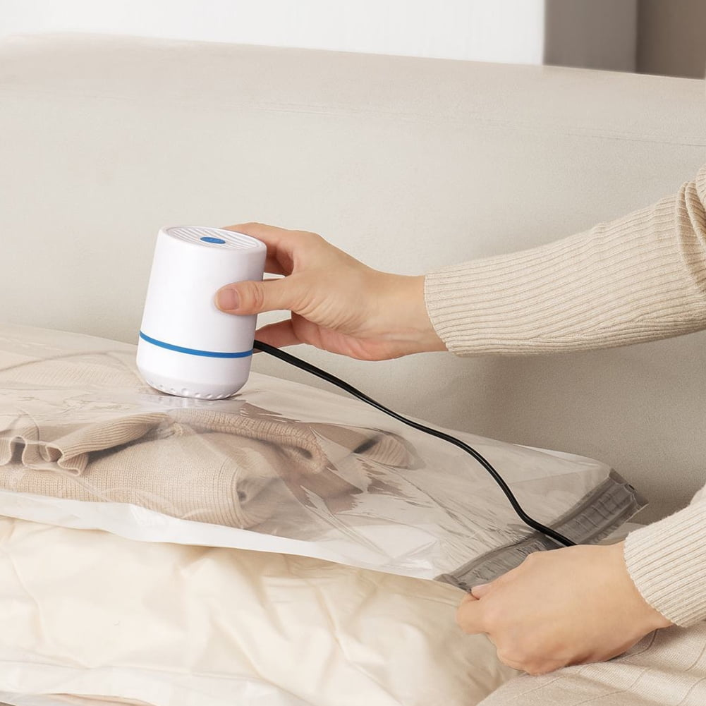 Eine Person verwendet einen weiß-blauen Staubsauger, um Kleidung in einem transparenten Aufbewahrungsbeutel auf einem weißen Bett zu komprimieren.