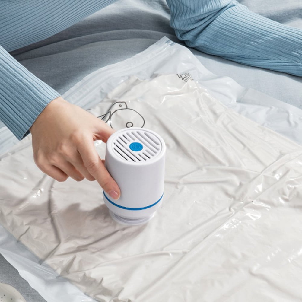 Eine Person benutzt einen weiß-blauen Staubsauger, um die Luft aus einem Aufbewahrungsbeutel mit Kleidung zu entfernen. Der Beutel liegt auf einem Bett.