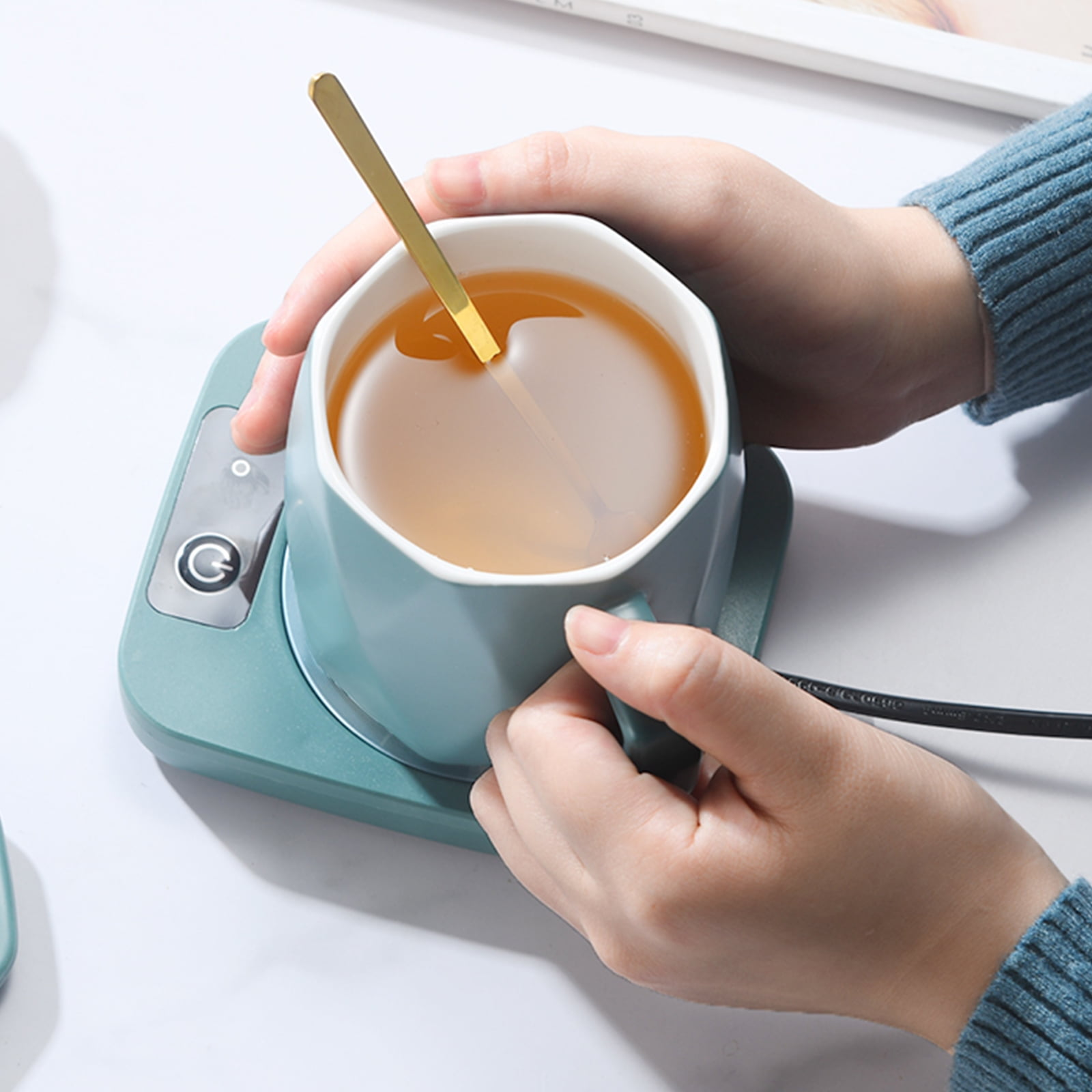 Hände halten eine türkisfarbene Tasse mit Tee und einem goldenen Löffel auf einem türkisfarbenen Wärmer. Die Person trägt einen blauen Pullover.