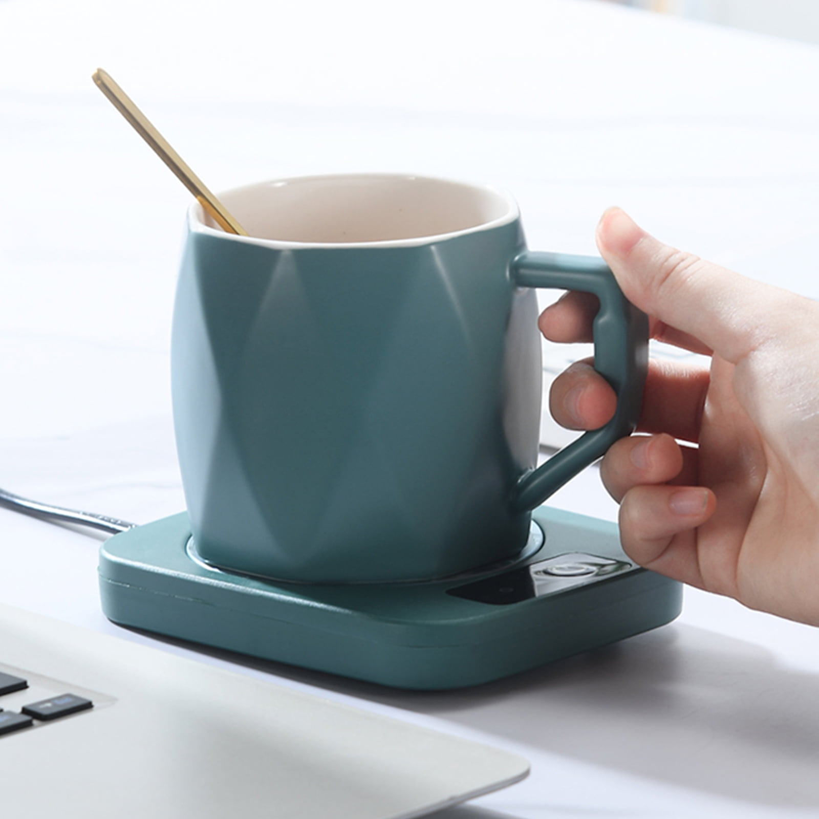 Eine türkisfarbene Tasse mit einem goldenen Löffel steht auf einem türkisfarbenen Wärmer. Eine Hand hält den Griff der Tasse, in der Nähe eines Laptops.