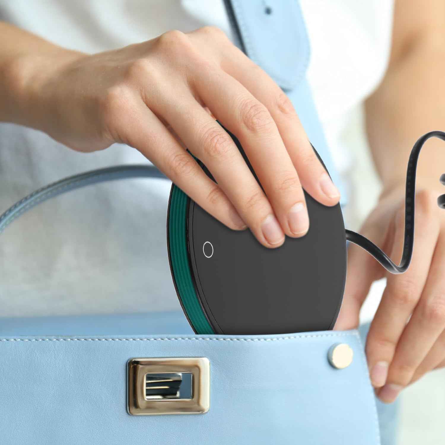 Eine Person legt ein schwarzes, kreisförmiges Ladegerät in eine hellblaue Handtasche. Das Ladegerät hat einen grünen Rand und einen weißen Einschaltknopf.