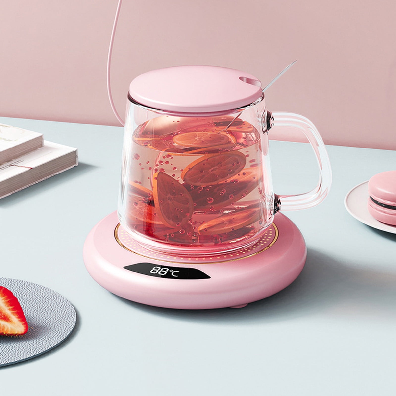 Ein rosa elektrischer Wasserkocher mit einer Glaskanne, gefüllt mit Tee und Zitronenscheiben, steht auf einem Tisch. Eine Erdbeerhälfte ist sichtbar.