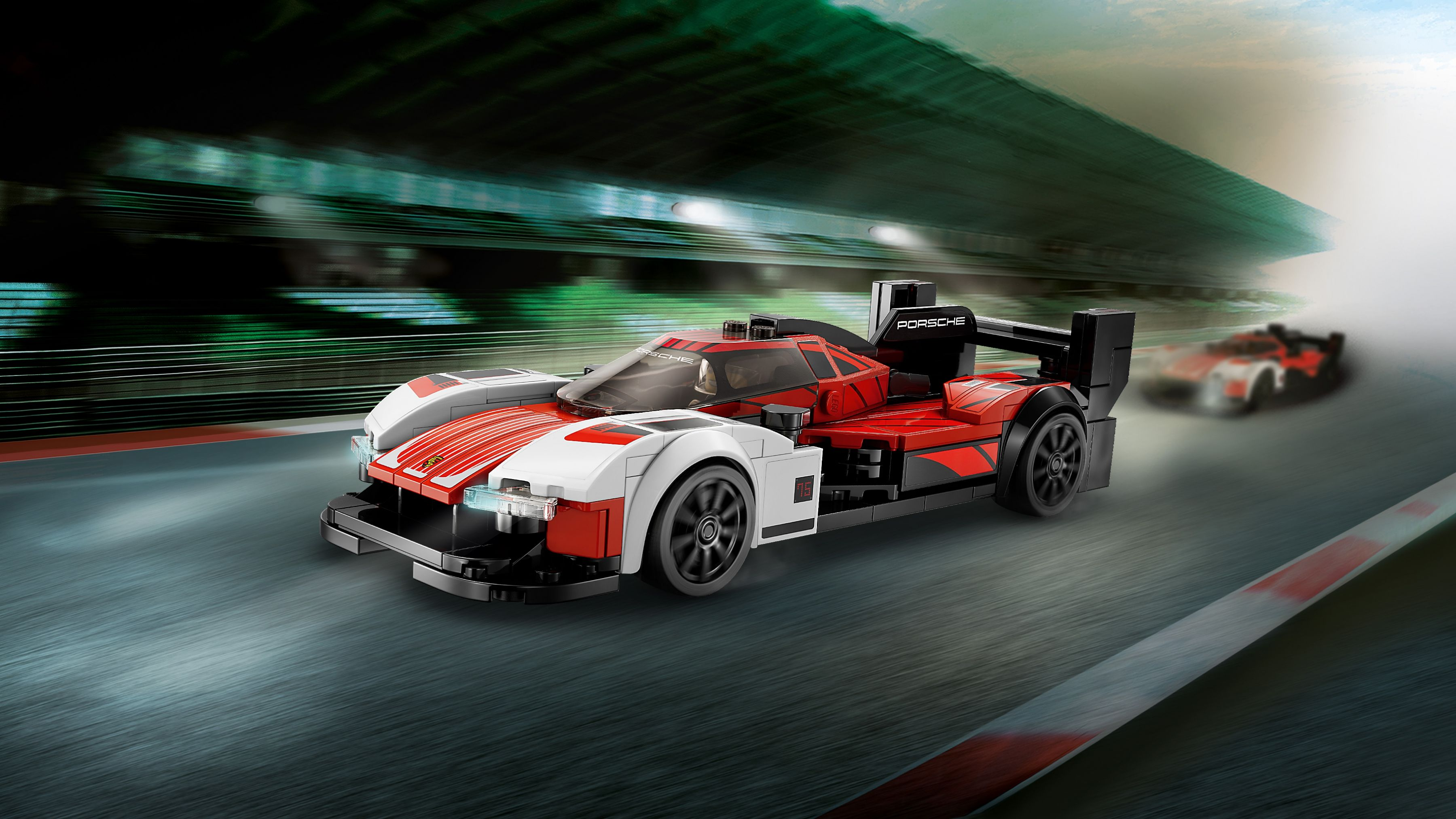 Ein rot-weißer Lego Porsche-Rennwagen auf einer Rennstrecke. Ein weiteres Auto verschwommen im Hintergrund, fährt. Die Szene ist schnell und dynamisch.