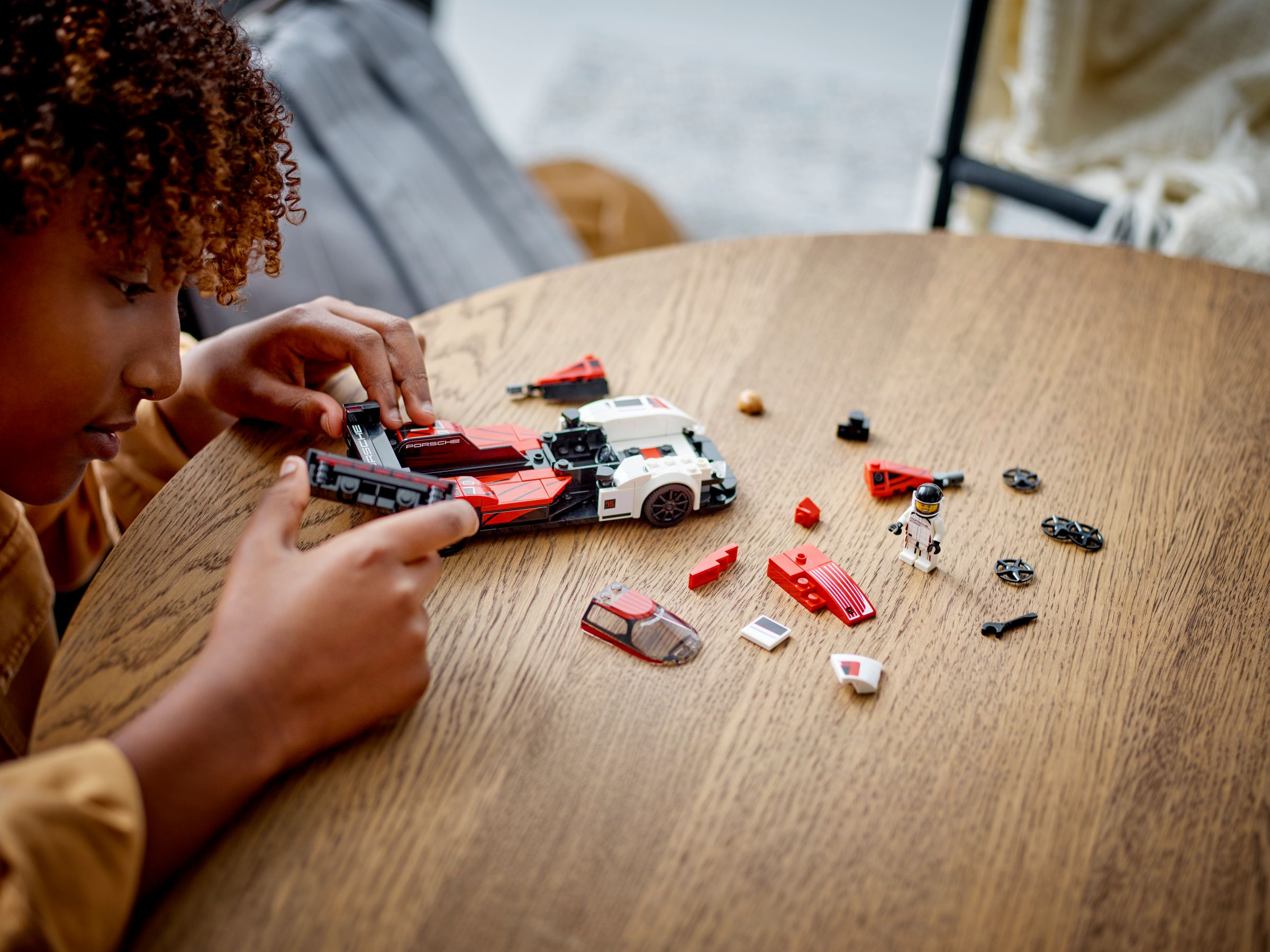 Eine Person baut ein rotes und weißes Lego Porsche-Auto auf einem Holztisch zusammen, mit verstreuten Teilen.