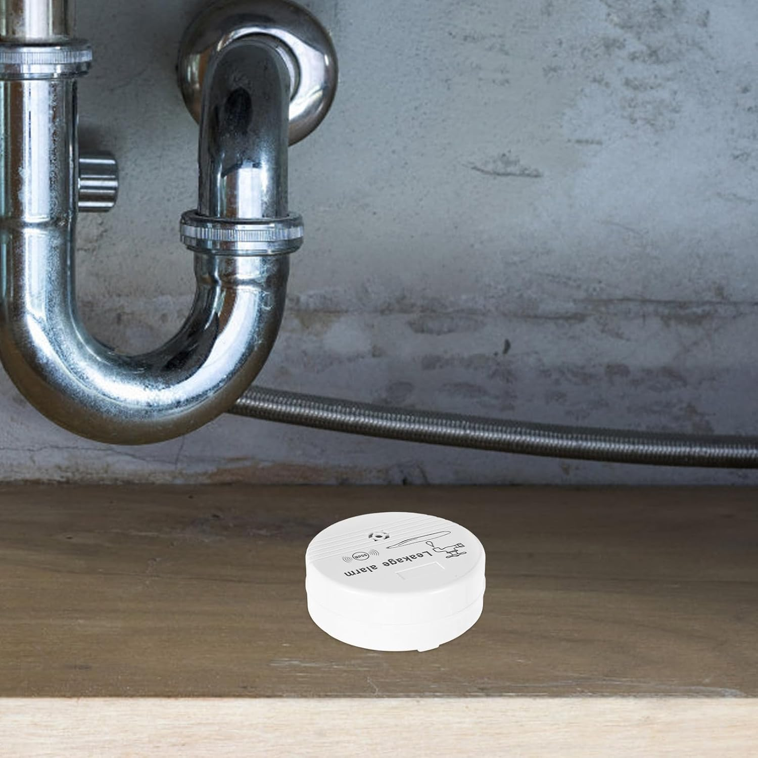 Ein weißer Leckalarm steht auf einem Holzregal unter Sanitärrohren. Der Hintergrund ist eine graue Betonwand.