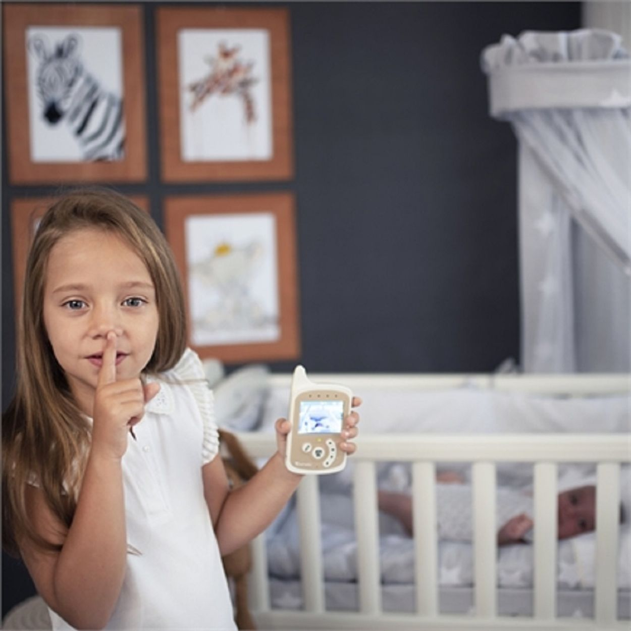 Ein Mädchen hält einen Babyphone und deutet auf Stille. Ein Baby schläft in einem Kinderbett. Kunstwerke hängen an der Wand.