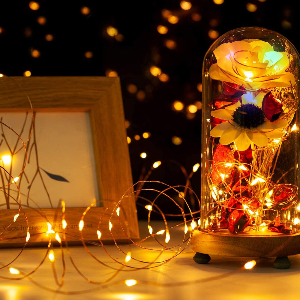 Eine dekorative Glaskuppel mit Blumenarrangement und Lichterketten steht neben einem gerahmten Bild. Weiche Bokeh-Lichter im Hintergrund.