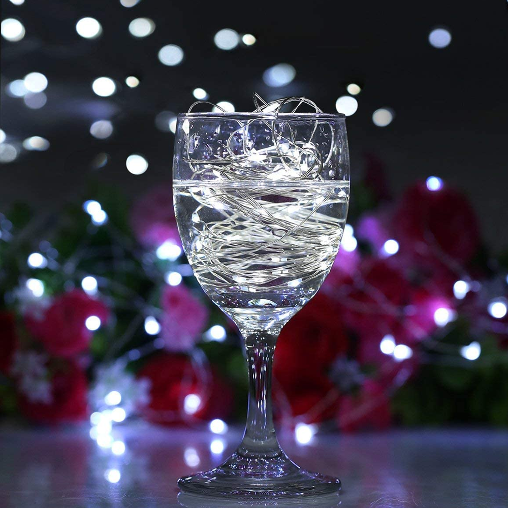 Ein Weinglas mit Wasser und Lichterketten gefüllt. Im Hintergrund sind rote Blumen und Bokeh-Lichter zu sehen.