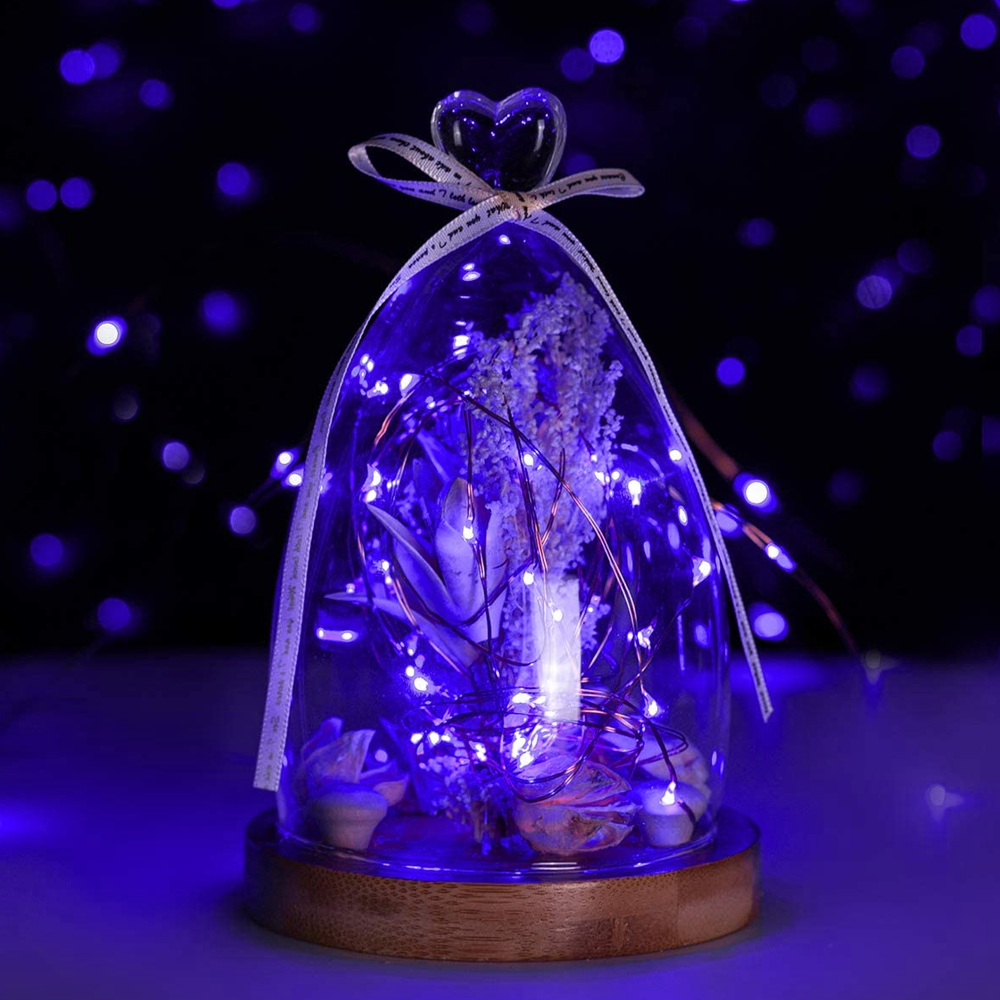 Glaskuppel mit Lichtern, herzförmige Spitze und Blumengesteck. Holzsockel, lila Lichter, dunkler, verschwommener Hintergrund.