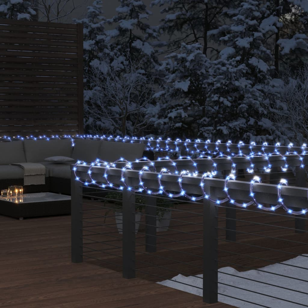 Eine nächtliche Szene im Freien zeigt eine Terrasse mit blauen Lichterketten. Im Hintergrund sind schneebedeckte Bäume zu sehen.