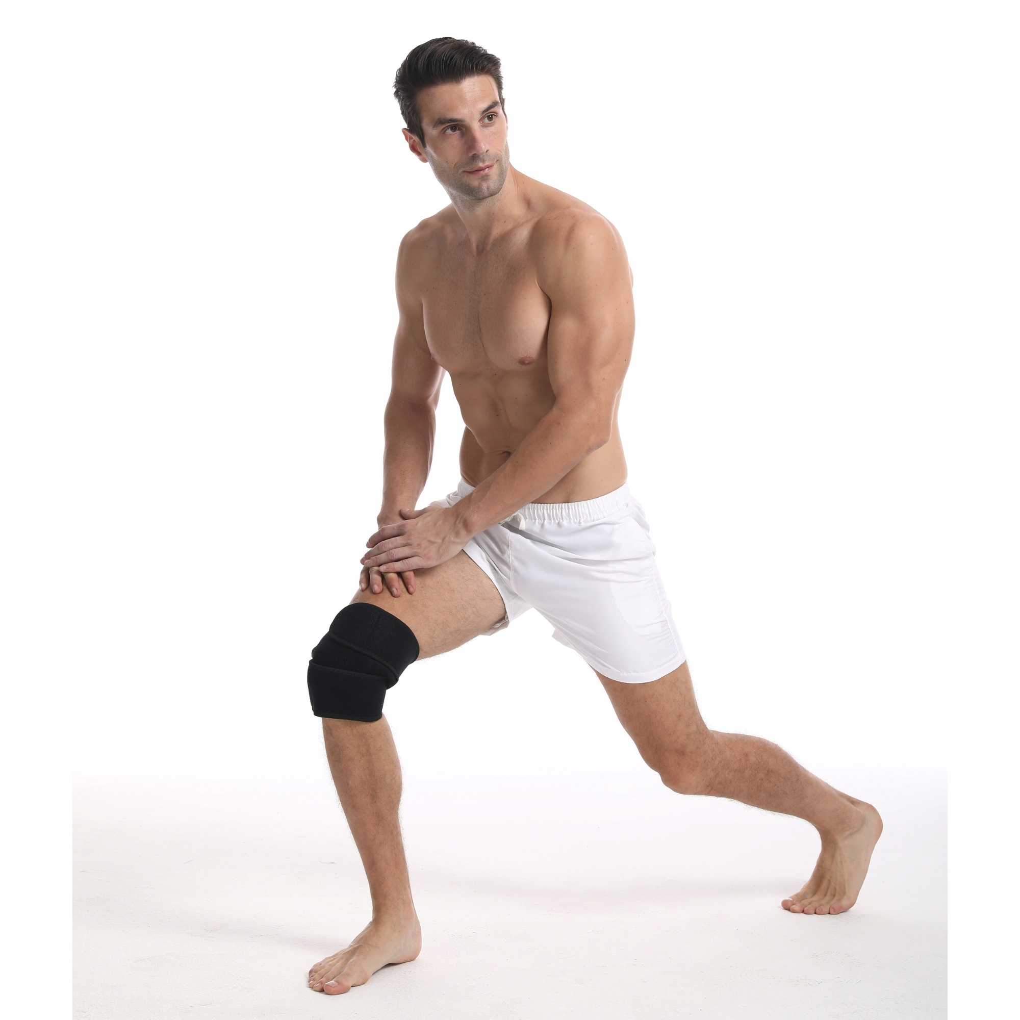 Mężczyzna w białych szortach, z orteza kolana, rozciąga się. Jest bez koszuli. Białe tło.