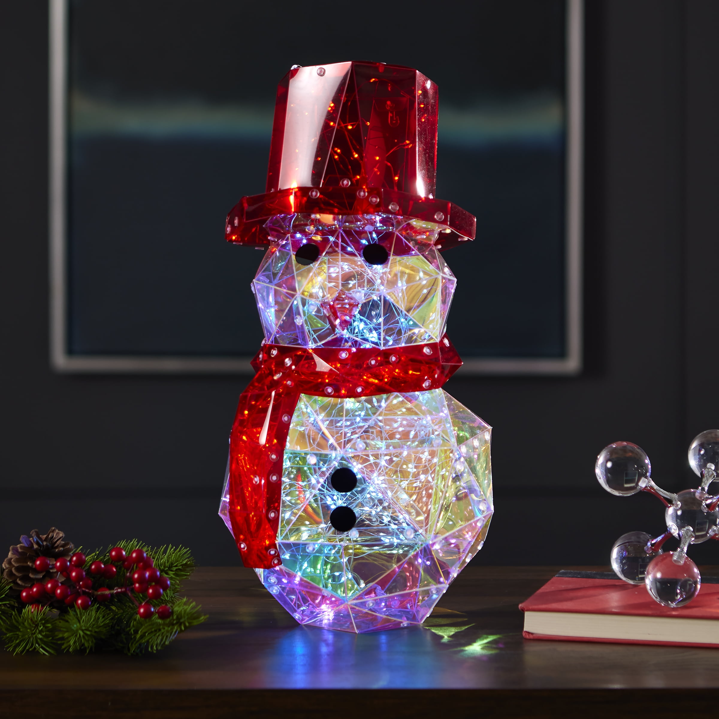 Ein Kristall-Schneemann mit rotem Hut und Schal, der mit bunten Lichtern beleuchtet ist. Er steht auf einem Tisch mit Dekorationen.