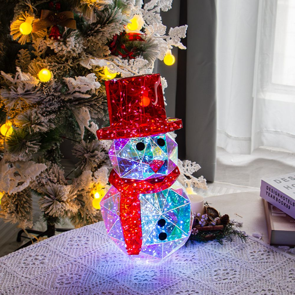 Ein leuchtender geometrischer Schneemann mit rotem Hut und Schal sitzt auf einem Tisch in der Nähe eines Weihnachtsbaums. Lichter beleuchten die Szene.