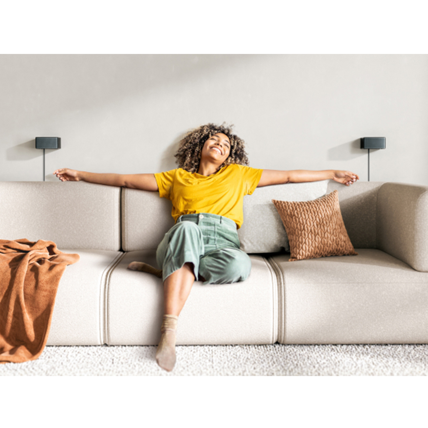 Kobieta w żółtej koszuli i zielonych spodniach siedzi na beżowej sofie, ramiona wyciągnięte. Brązowy koc i poduszka, głośniki na ścianie.