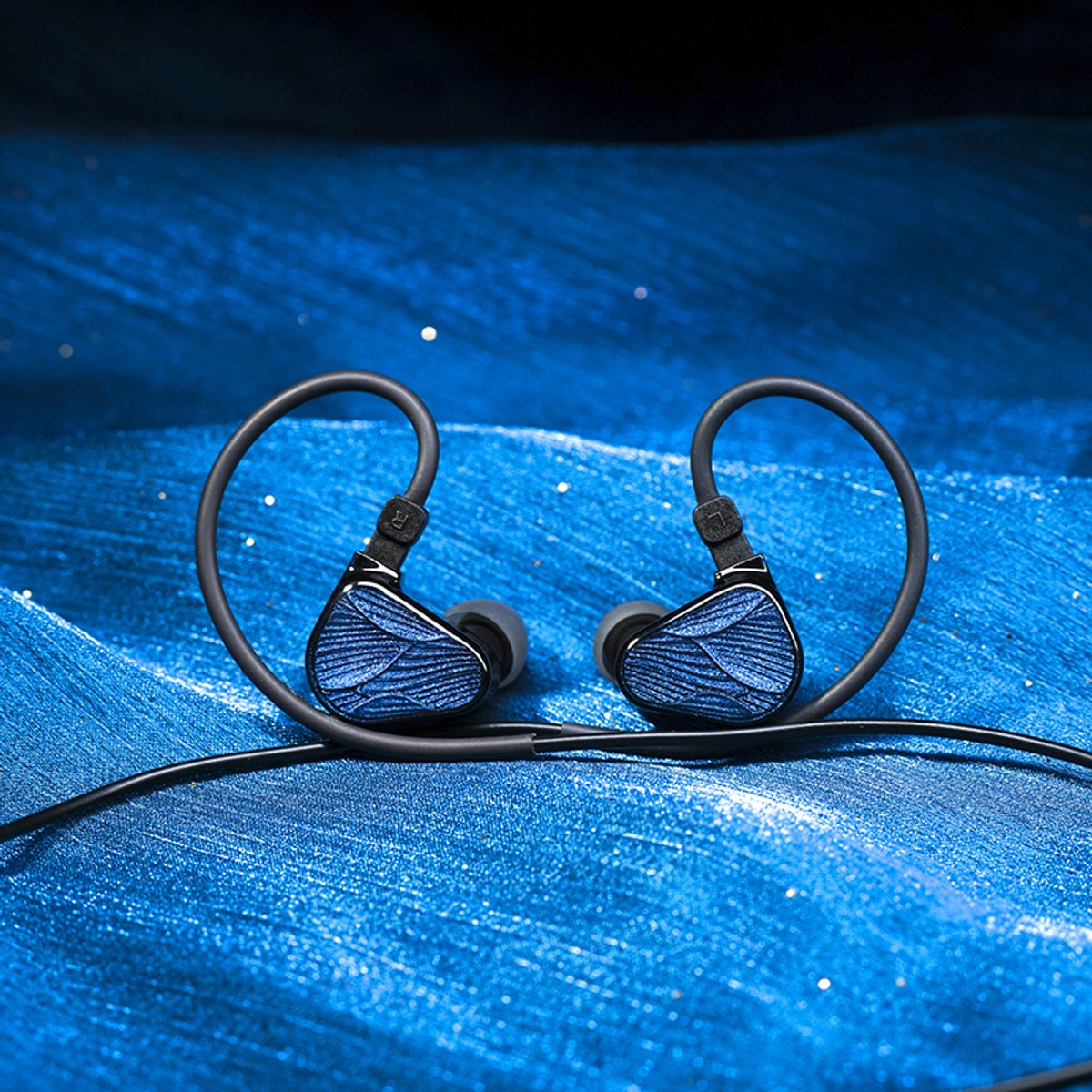 Zwei blaue und schwarze In-Ear-Kopfhörer auf einer blau strukturierten Oberfläche. Der Hintergrund ist ebenfalls blau mit weißen Punkten.