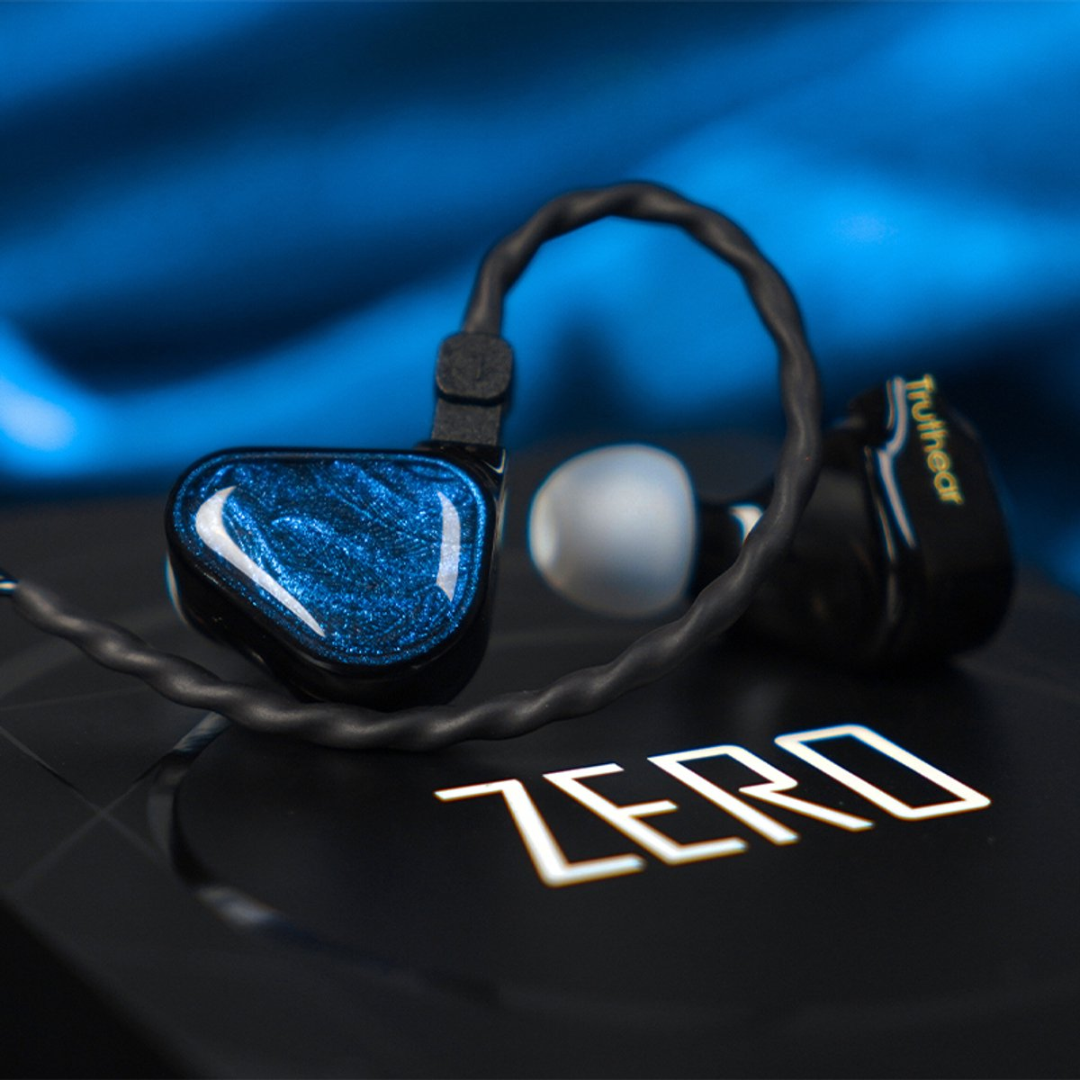 Nahaufnahme von schwarzen und blauen Ohrhörern mit Kabel, das Wort ZERO und der Markenname Truhear sind sichtbar. Der Hintergrund ist blau.