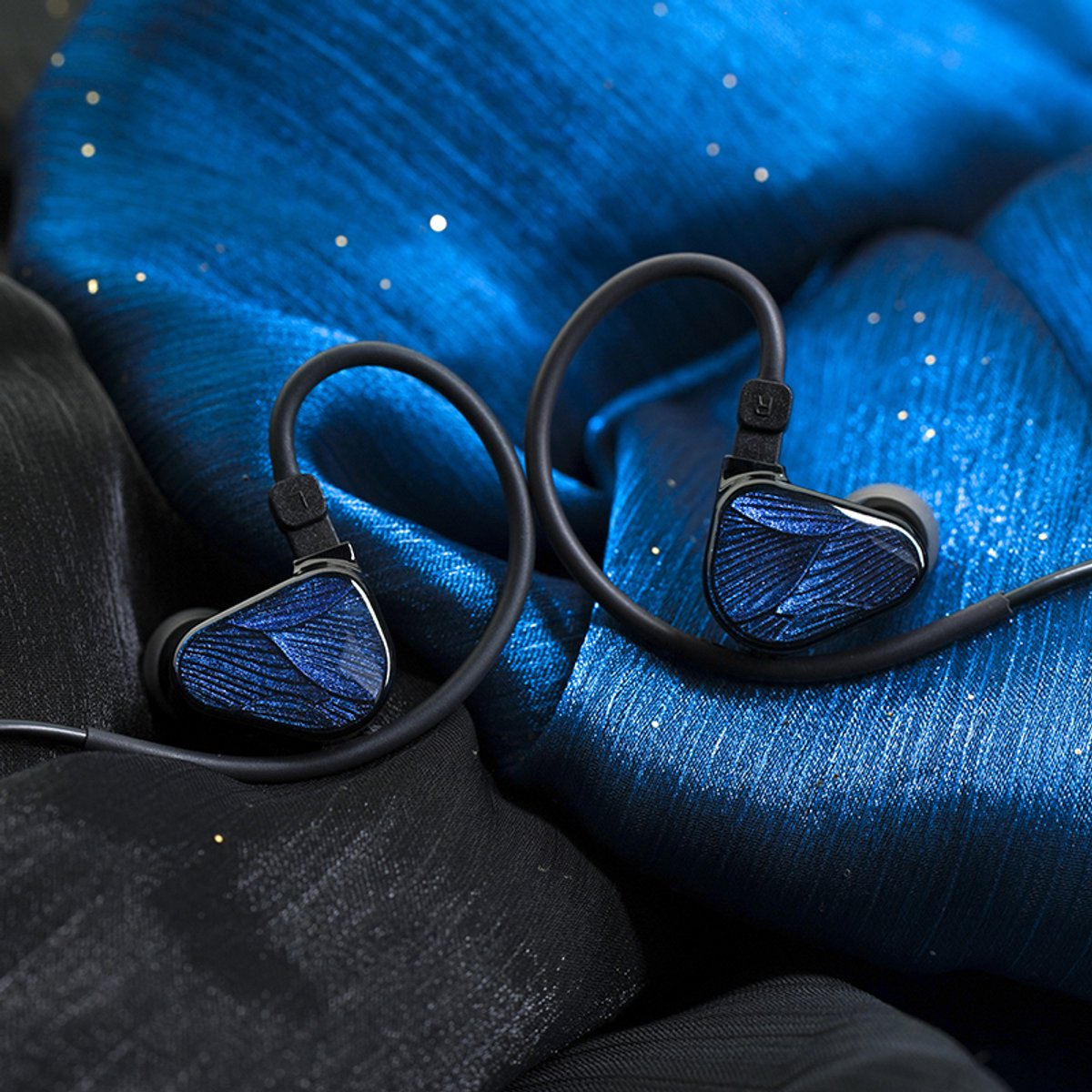 Zwei In-Ear-Kopfhörer mit blau gemusterten Flächen liegen auf einem strukturierten, blauen und schwarzen Stoffhintergrund.