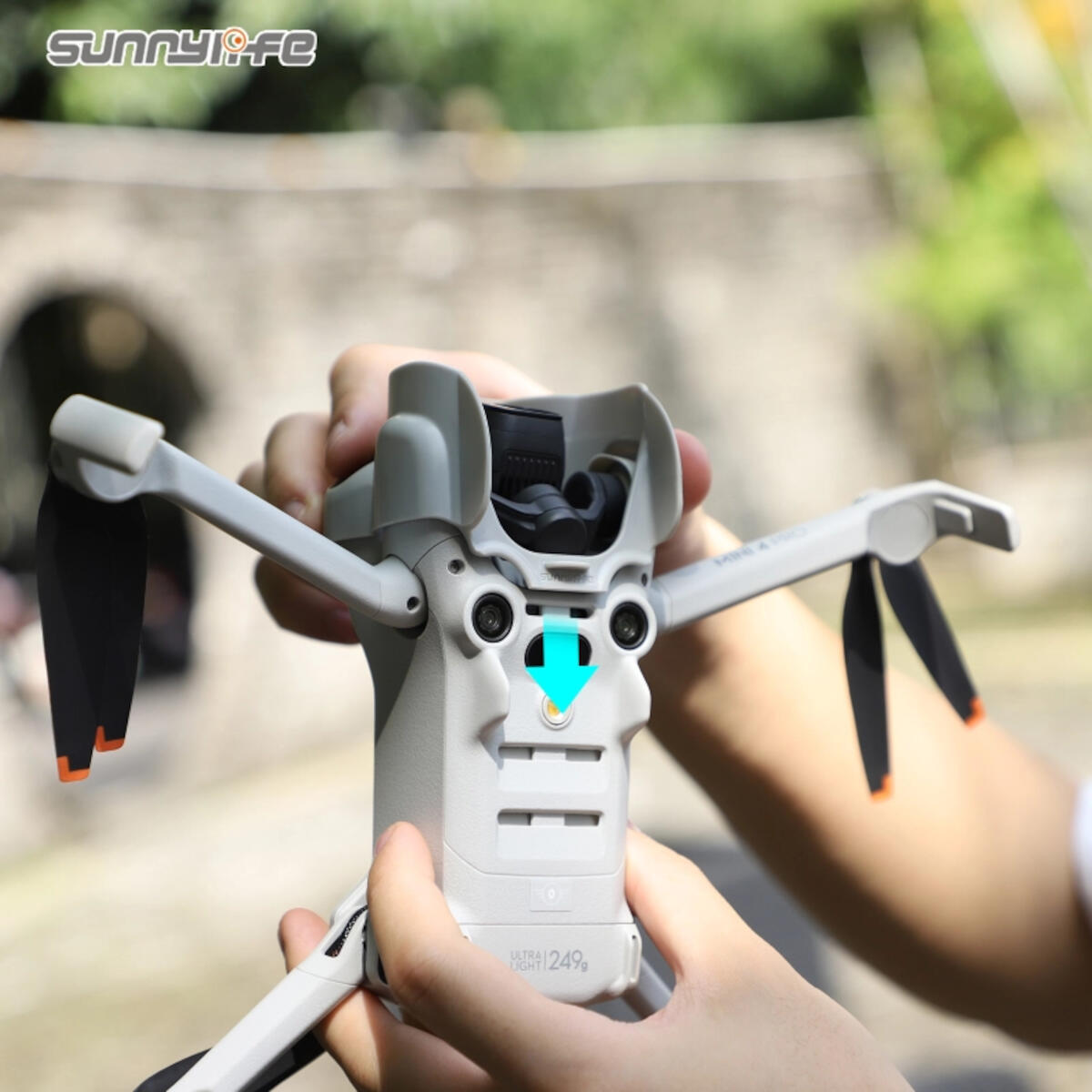 A person holds a white drone with black propellers. The drone has an orange arrow indicating a button. Green background.