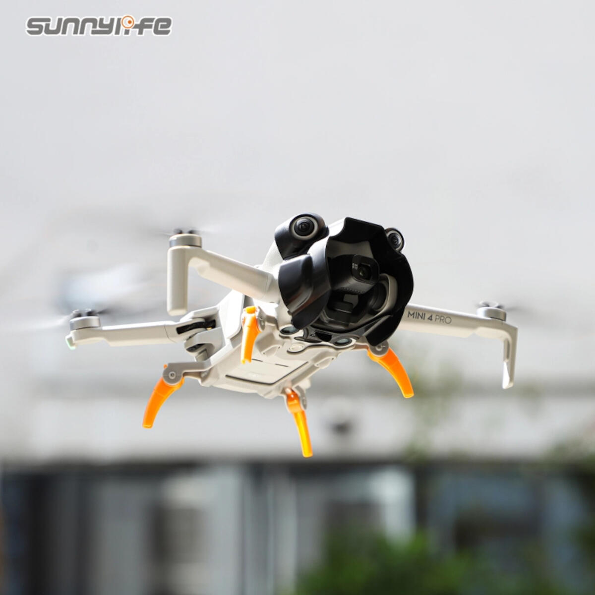 A white drone with black camera hood and orange landing gear is in flight. The drone has four propellers and the text 'MINI 4 PRO'.