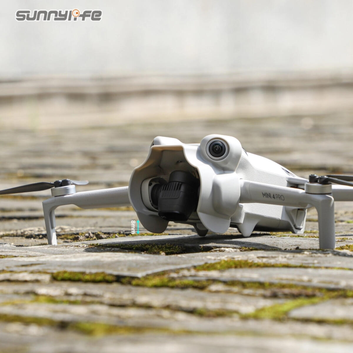 A gray drone, 'MINI 4 PRO', sits on a stone surface. The drone has a camera and propellers. The background is a blurred stone path.