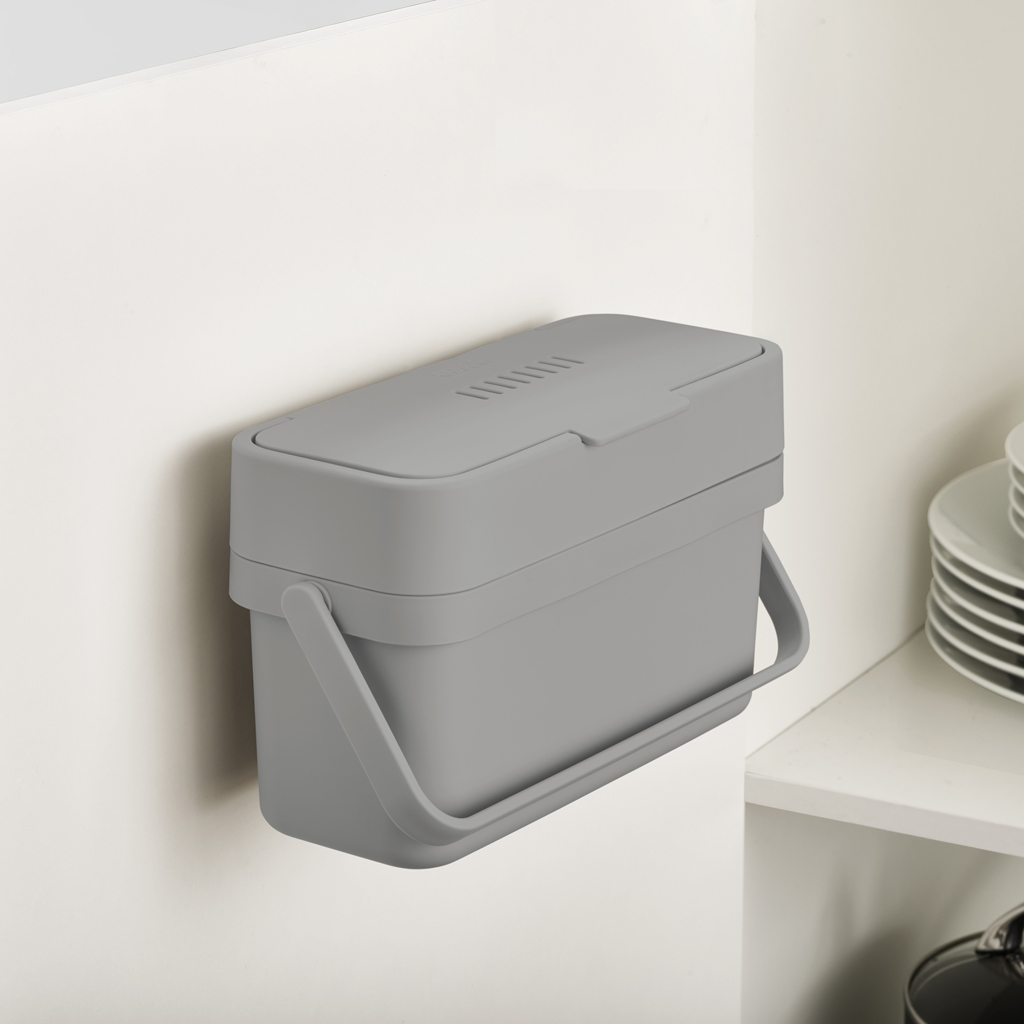 Un cubo de compostaje gris con asa está montado en una pared blanca. Una pila de platos blancos está sobre un estante.