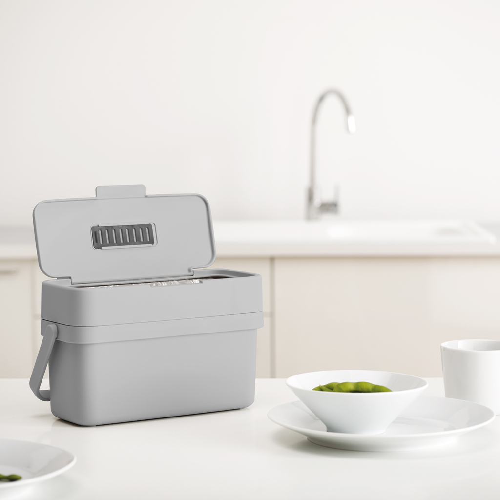 Un contenedor de compostaje gris con la tapa abierta está sobre una mesa blanca. Un cuenco de judías verdes y una taza blanca también están sobre la mesa.