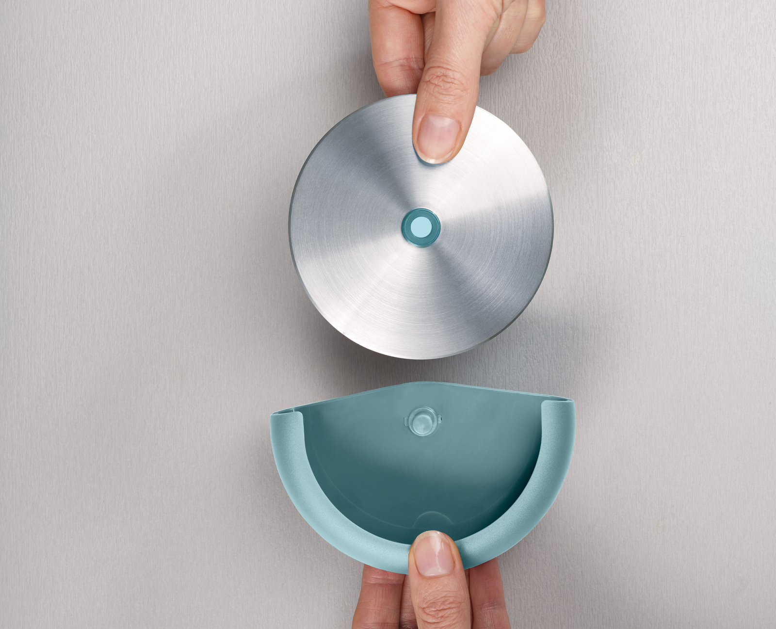 Manos sosteniendo una tapa plateada y un recipiente verde azulado. La tapa es redonda y el recipiente tiene forma de media luna.