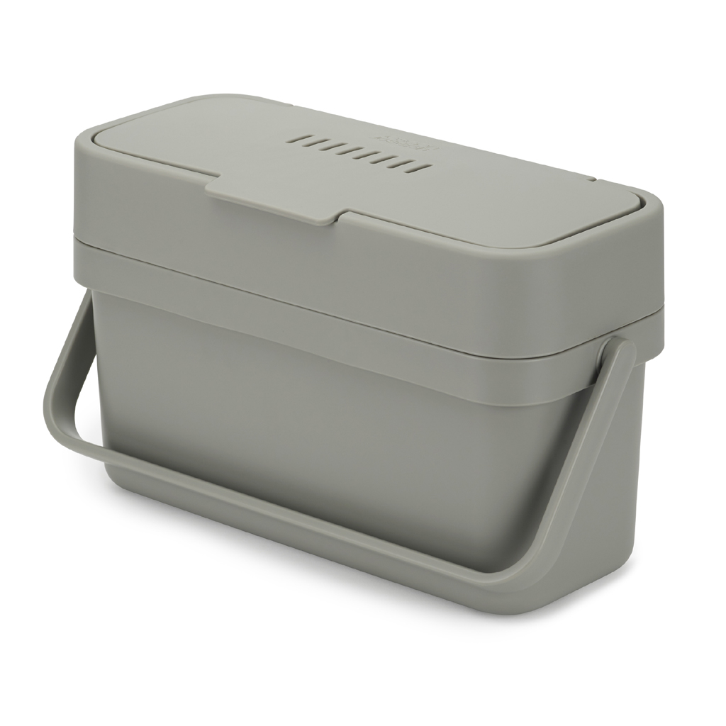 Un contenedor de compostaje rectangular gris con tapa y asa. La tapa tiene ventilas. Se encuentra sobre una superficie blanca.