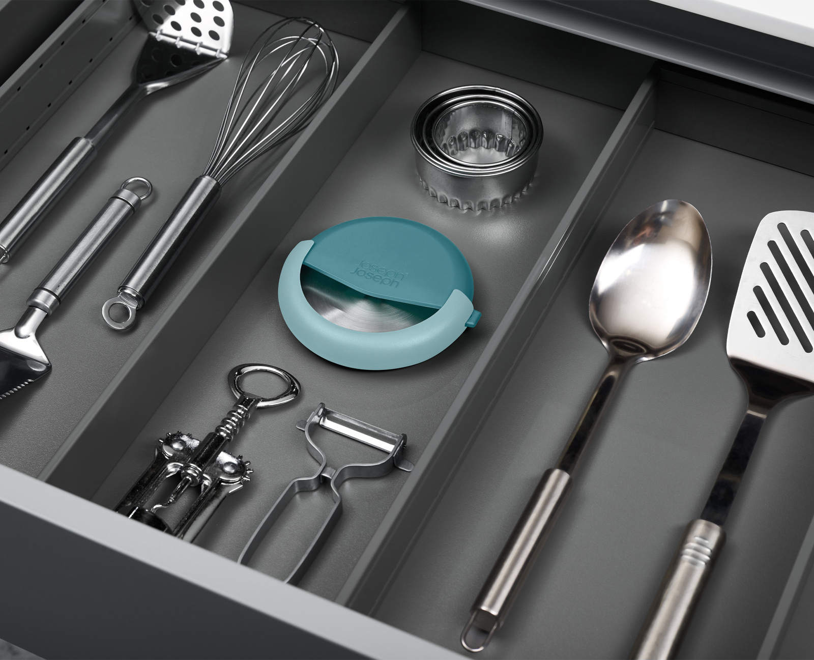 Un cajón con varios utensilios de cocina: batidora, espátula, cuchara, pelador, abridor de botellas y un cortador de pizza azul.