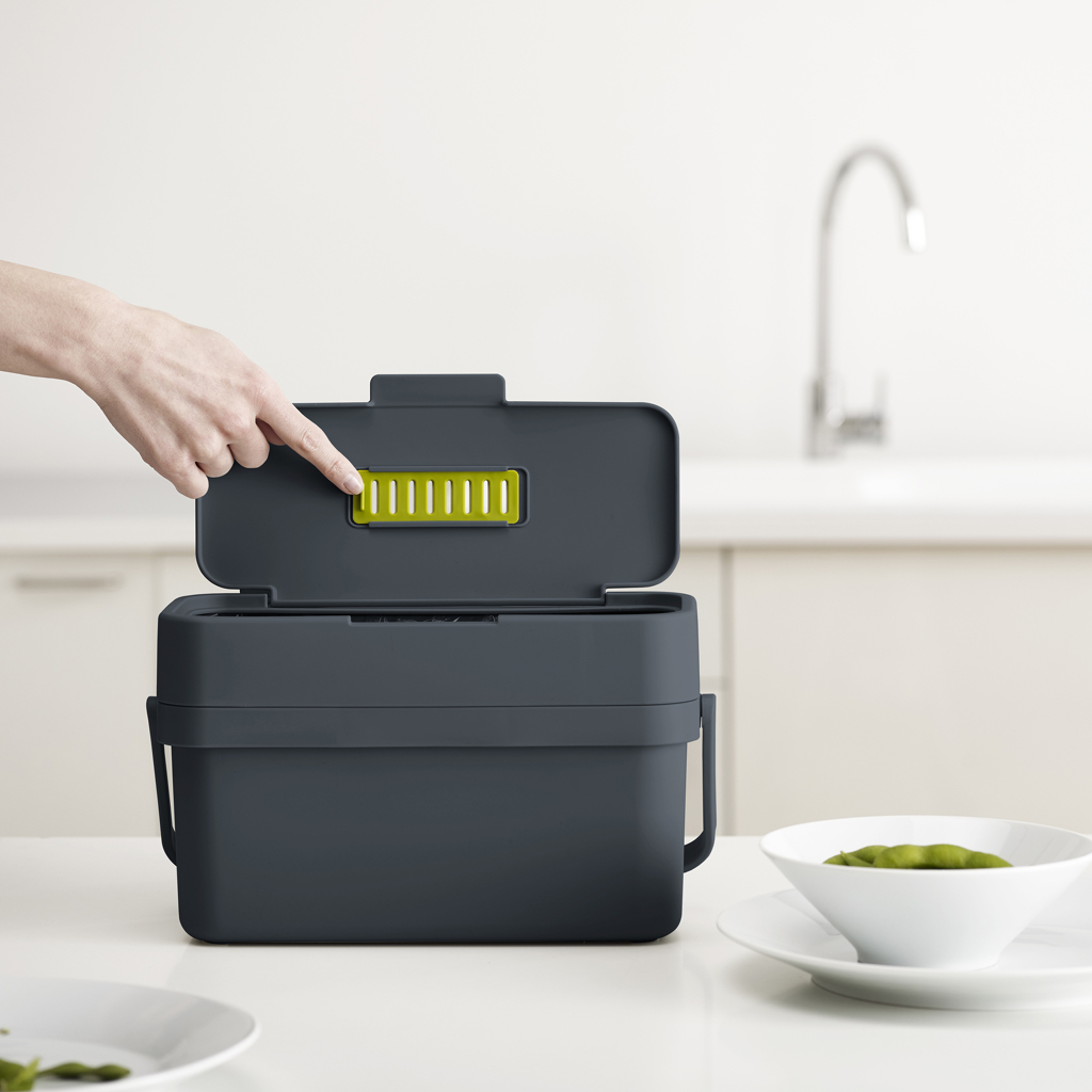 Una mano abre un cubo de compostaje gris oscuro. Un cuenco blanco con judías verdes está sobre un plato blanco. Hay un fondo de cocina.