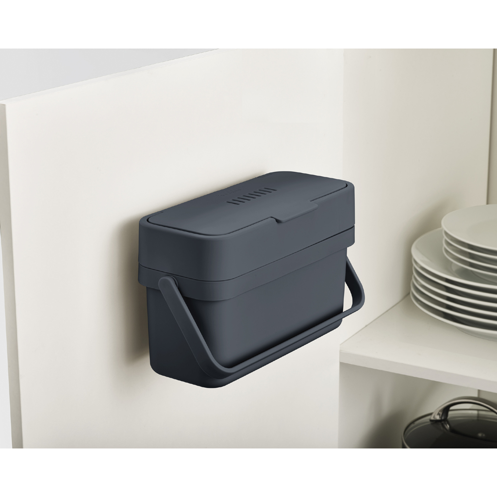 Un cubo de compostaje rectangular gris oscuro con asa está montado en un armario blanco. Platos y una olla están en un estante.