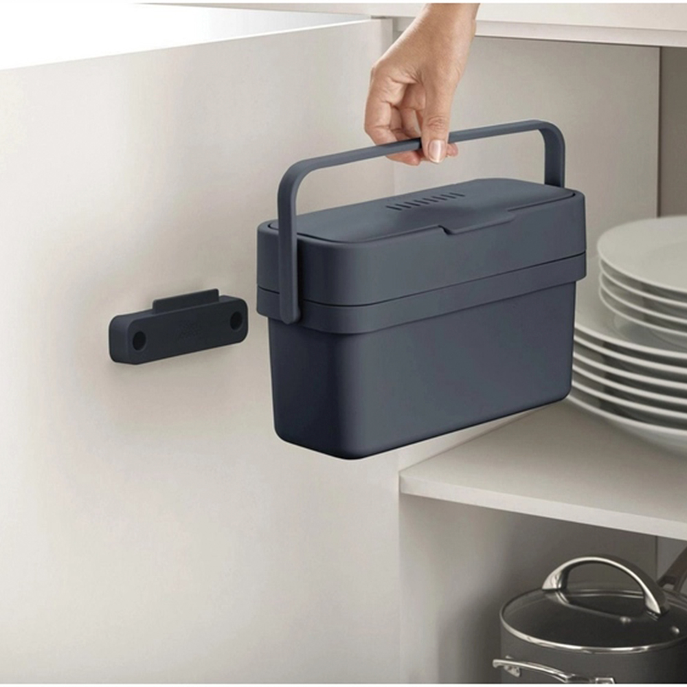 Una mano sostiene un contenedor azul oscuro con asa. Un soporte magnético negro está en la pared. Platos y una olla en el fondo.