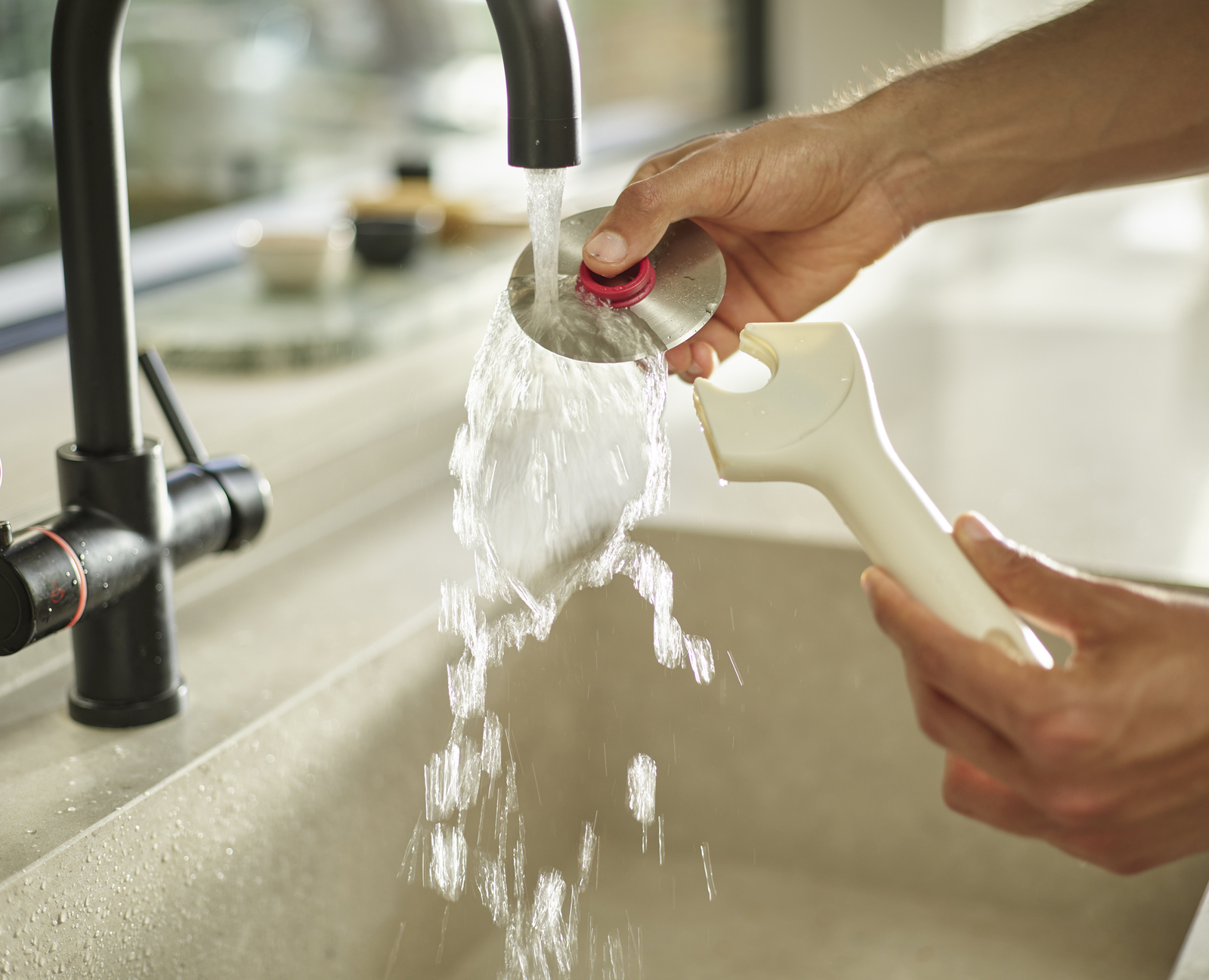 Manos lavando un objeto metálico bajo un grifo. Fluye agua, se sostiene un mango blanco. Se ven el fregadero y el grifo de la cocina.