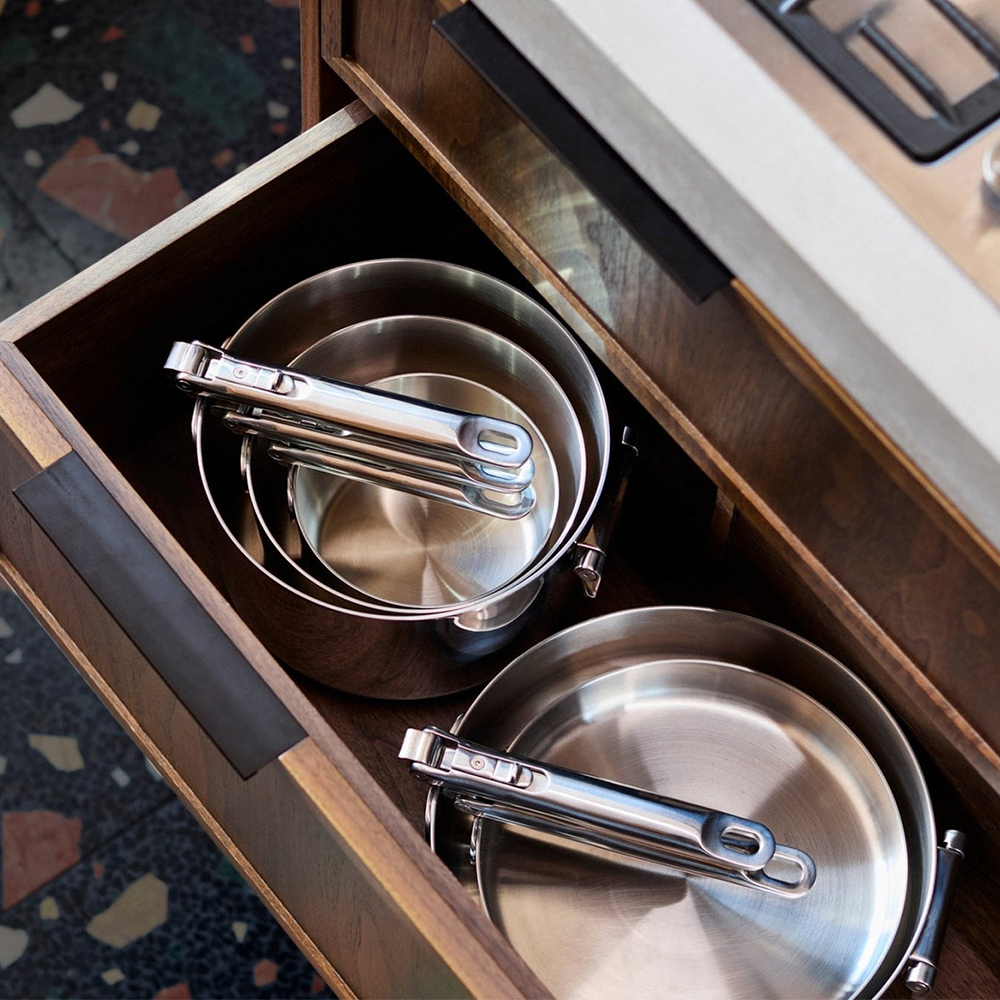 Ollas y sartenes de acero inoxidable con pinzas en un cajón de madera. Ambiente de cocina. Cajón parcialmente abierto.