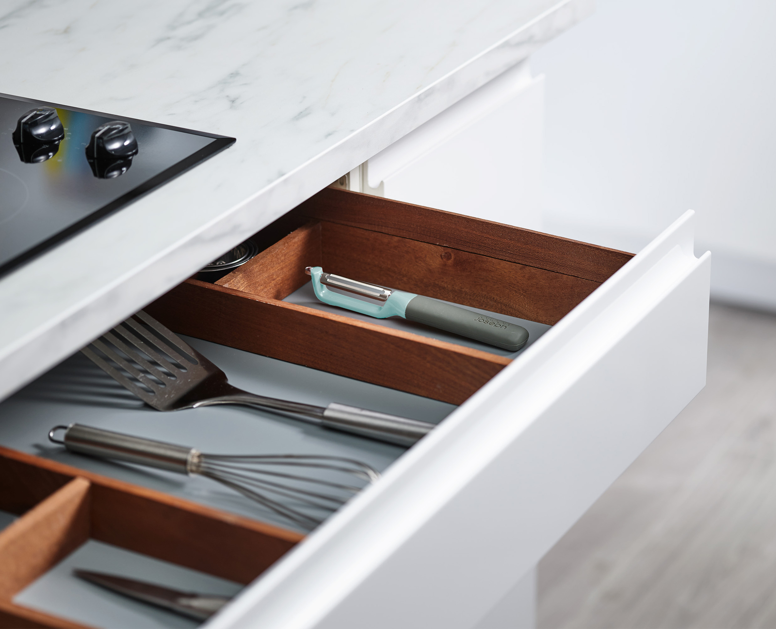 Cajón de cocina abierto con utensilios. Se ven un pelador, una espátula y un batidor. Encimera de mármol y una placa de cocina.