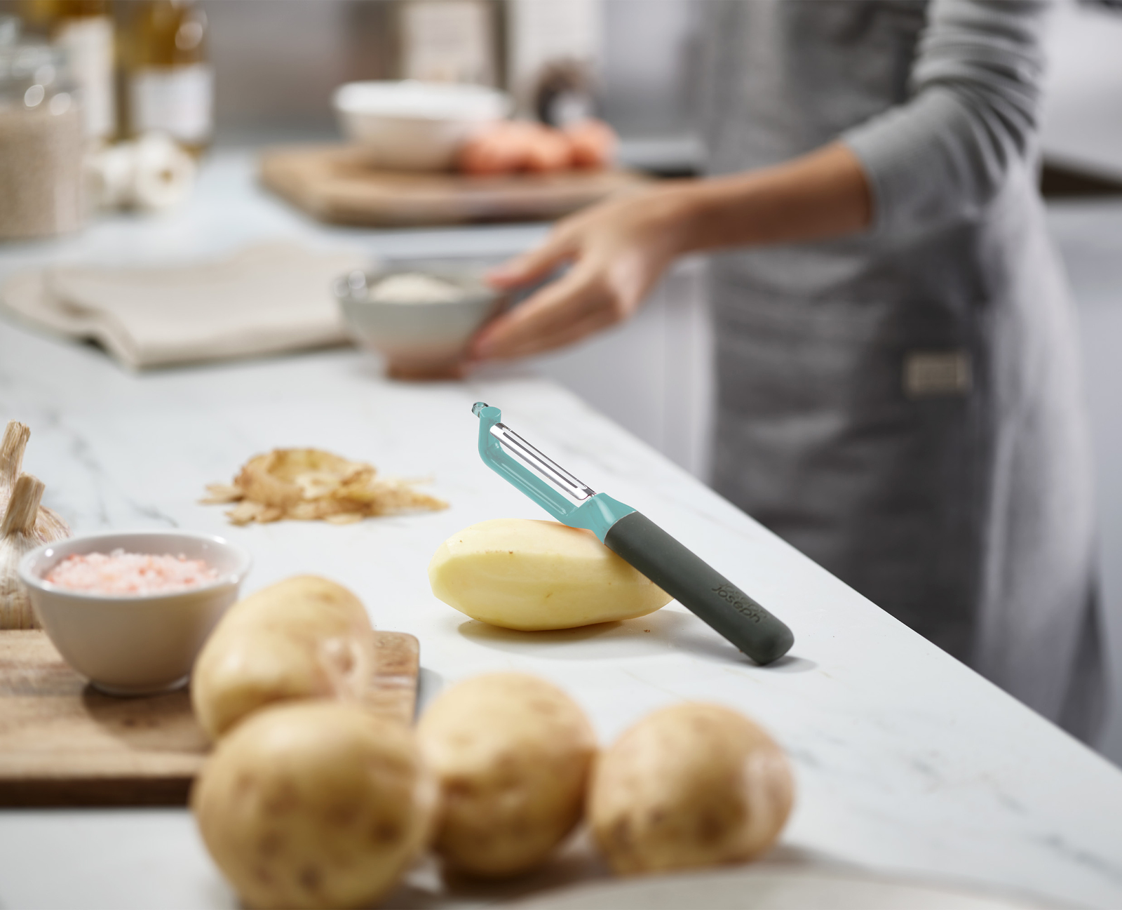 Una escena de cocina con un pelador, patatas y una persona con delantal. Se pela una patata en una encimera blanca.