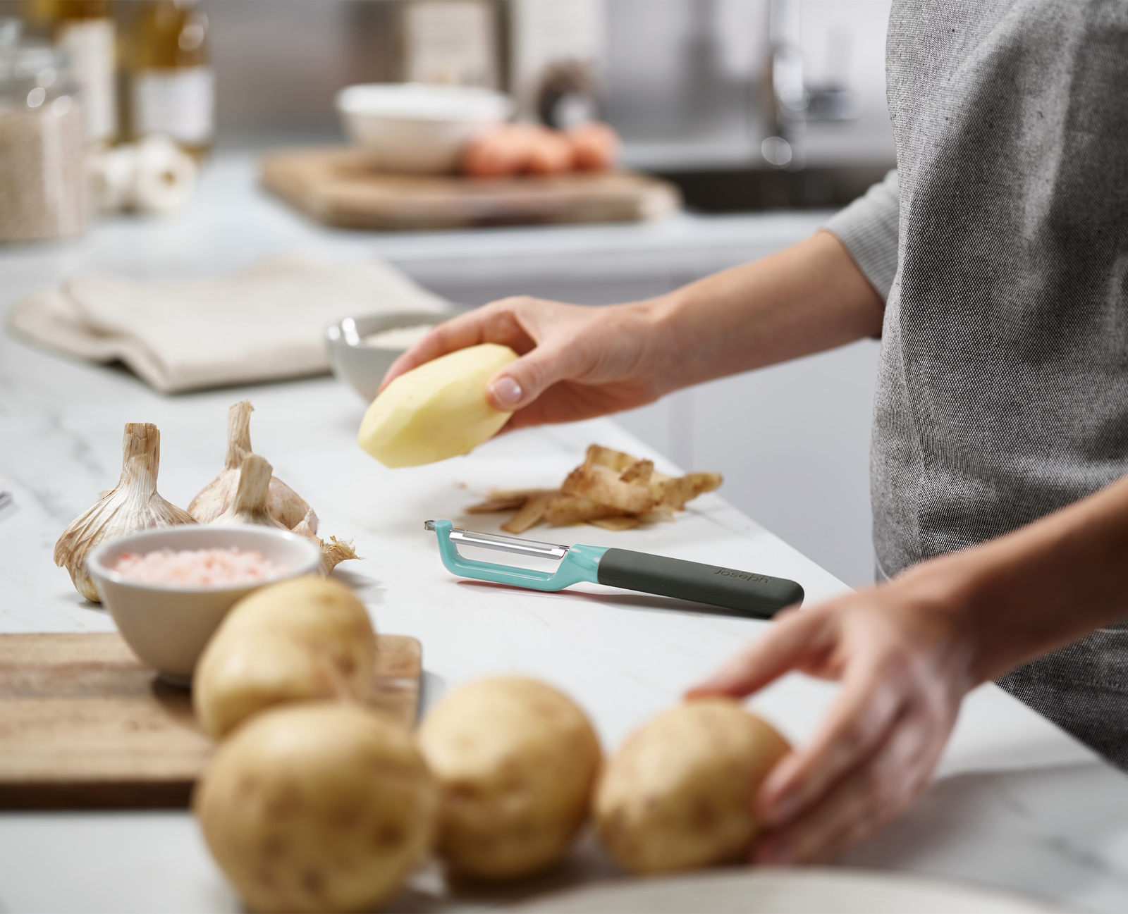 Una persona pela una patata con un pelador. Ajo, sal y patatas sobre una superficie blanca. Ambiente de cocina.