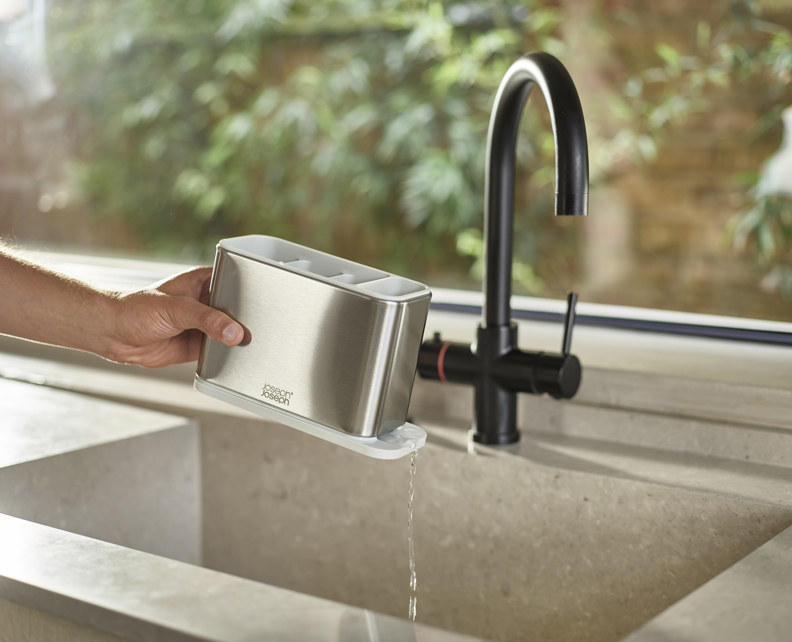 Una mano sostiene un porta utensilios de acero inoxidable sobre un fregadero. El agua fluye del soporte. Un grifo negro está en el fondo.