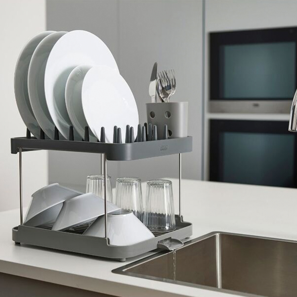 Un escurreplatos gris de dos niveles sostiene platos, cuencos y vasos blancos. Los utensilios están en un soporte gris. Un fregadero está en primer plano.