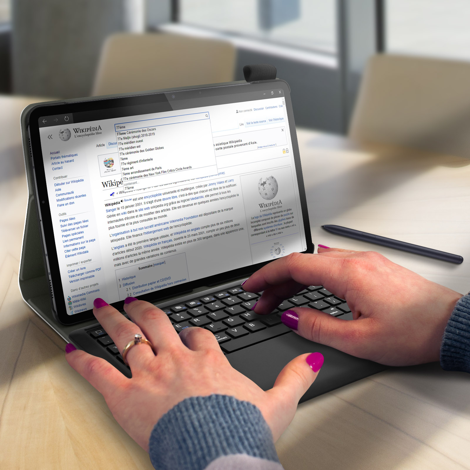 Ein Laptop mit Wikipedia-Seite auf einem Schreibtisch. Hände mit pinken Nägeln tippen.